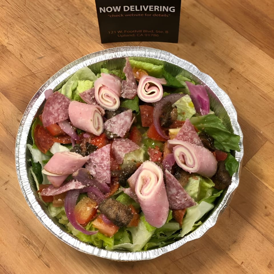 Antipasto salad with ham, salami, pepperoni, lettuce, red onion, and tomatoes in a foil container.